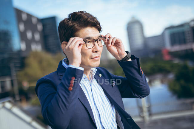 Porträt eines lächelnden asiatischen Geschäftsmannes, der seine Brille in einer Straße mit Gebäuden hinter sich berührt. Geschäftsmann im Stadtkonzept unterwegs. — Stockfoto