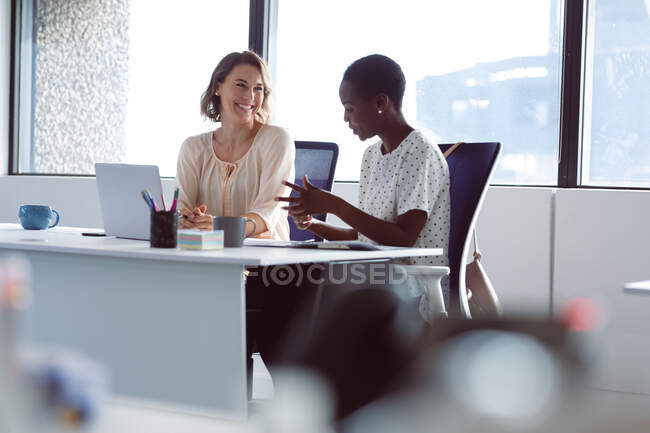 Dos empresarias diversas sentadas en el escritorio, hablando y sonriendo. negocio creativo independiente en una oficina moderna. - foto de stock