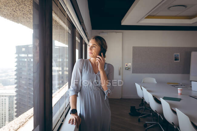 Kaukasische Geschäftsfrau, die am Fenster steht und bei der Arbeit mit dem Smartphone spricht. unabhängiges kreatives Geschäft in einem modernen Büro. — Stockfoto