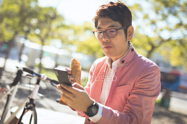 Hombre de negocios asiático usando smartphone comiendo bocadillo para llevar sentado en la pared en la ciudad. nómada digital en concepto de ciudad. - foto de stock