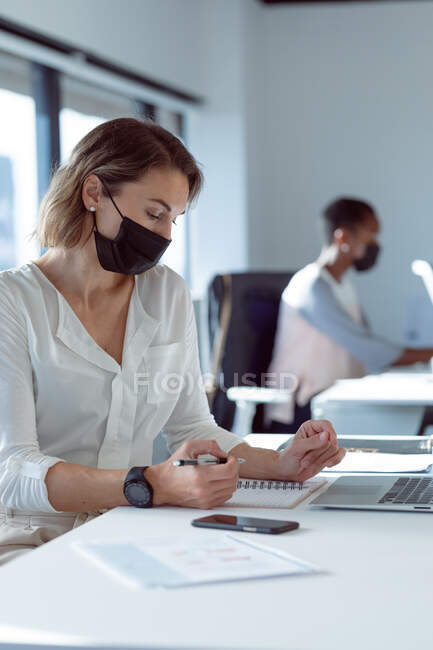 Mulher de negócios caucasiana a usar máscara facial, sentada à secretária, a fazer anotações. negócio criativo independente em um escritório moderno durante coronavírus covid 19 pandemia. — Fotografia de Stock