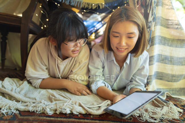Smiling asian woman with her daughter using tablet lying under tent in living room. at home in isolation during quarantine lockdown. — Stock Photo