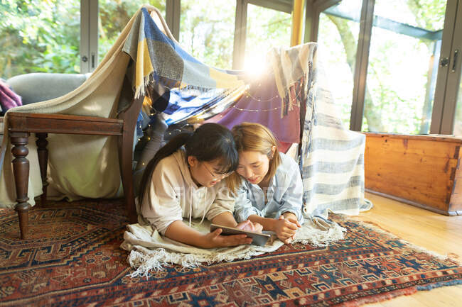 Sorrindo mulher asiática com sua filha usando tablet deitado sob tenda cobertor na sala de estar. em casa em isolamento durante o confinamento de quarentena. — Fotografia de Stock