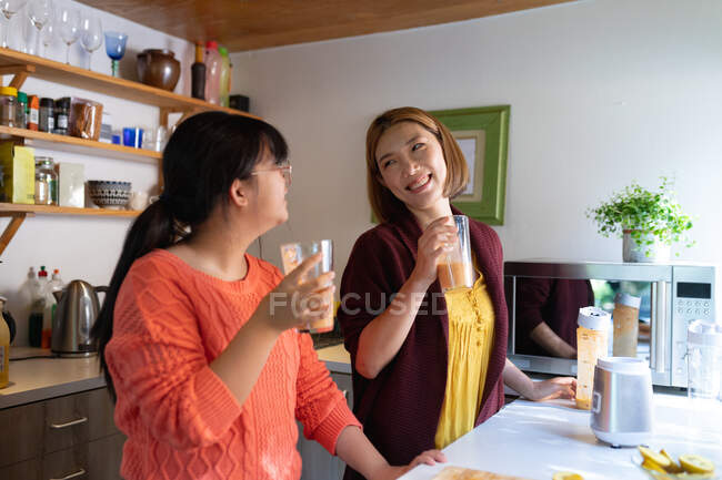 Sonriente asiática madre e hija bebiendo batido casero juntos en la cocina. en casa en aislamiento durante el bloqueo de cuarentena. - foto de stock