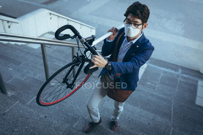 Asian businessman wearing face mask carrying bike up steps in city street. businessman out and about in city during covid 19 pandemic concept. — Stock Photo
