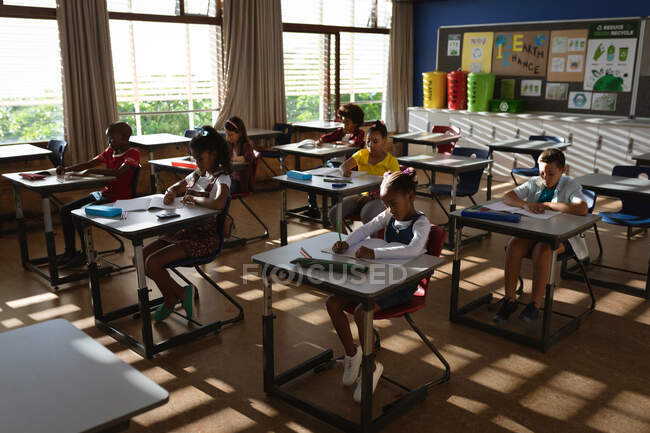 Diverso grupo de estudiantes que estudian mientras están sentados en su escritorio en clase en la escuela primaria. escuela y concepto de educación - foto de stock