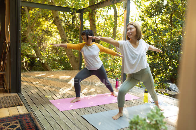 Улыбающаяся азиатка и ее дочь практикуют йогу на террасе в саду. в доме в изоляции во время карантинной изоляции. — стоковое фото