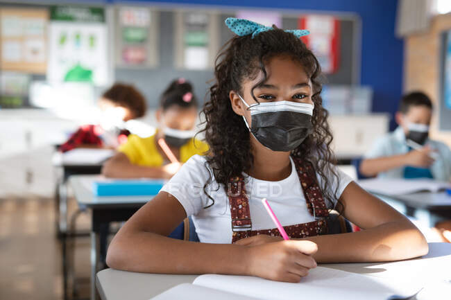 Retrato de menina afro-americana usando máscara facial sentada em sua mesa na escola primária. higiene e distanciamento social na escola durante a pandemia de 19 anos — Fotografia de Stock