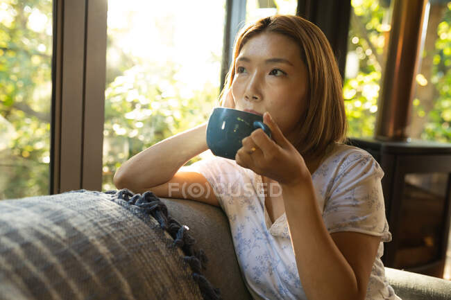 Porträt einer asiatischen Frau, die Tee trinkt und auf dem Sofa sitzt. Zuhause in Isolation während der Quarantäne. — Stockfoto