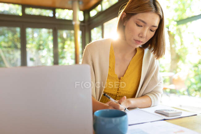 Asiatin macht sich Notizen, sitzt am Tisch und arbeitet von zu Hause aus. Zuhause in Isolation während der Quarantäne. — Stockfoto