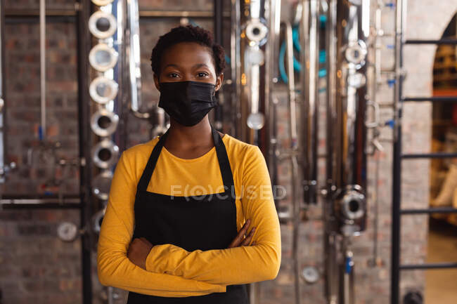 Портрет африканской американской работницы в маске для лица, стоящей на заводе по производству джина. Концепция производства и фильтрации алкоголя — стоковое фото
