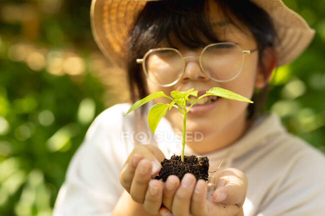 Sorrindo menina asiática vestindo óculos e chapéu de palha, segurando planta no jardim. em casa em isolamento durante o confinamento de quarentena. — Fotografia de Stock