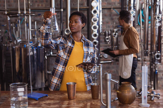 Trabalhadora afro-americana segurando um balão e um tablet digital na destilaria gin. conceito de produção e filtração de álcool — Fotografia de Stock