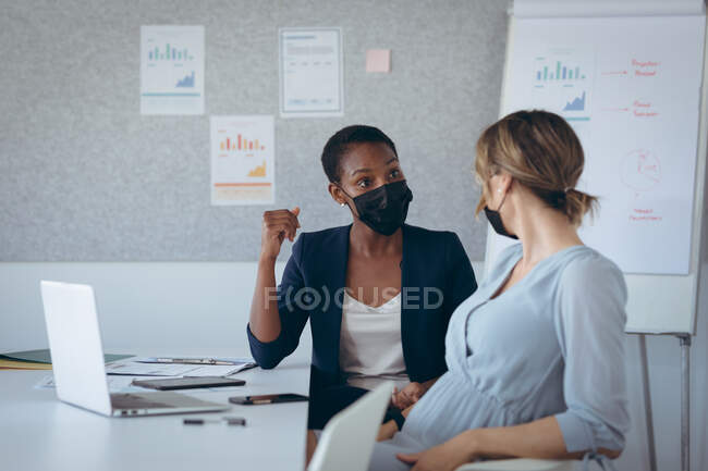 Two diverse businesswomen wearing face masks, sitting at desk, using laptop, talking. independent creative business at a modern office during coronavirus covid 19 pandemic. — Stock Photo