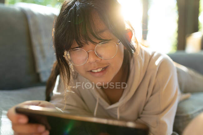 Sorrindo menina asiática em óculos usando smartphone e deitado no sofá. em casa em isolamento durante o confinamento de quarentena. — Fotografia de Stock