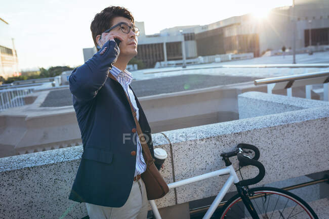 Hombre de negocios asiático hablando en un teléfono inteligente sosteniendo el café para llevar en su bicicleta en la calle de la ciudad. nómada digital en concepto de ciudad. - foto de stock