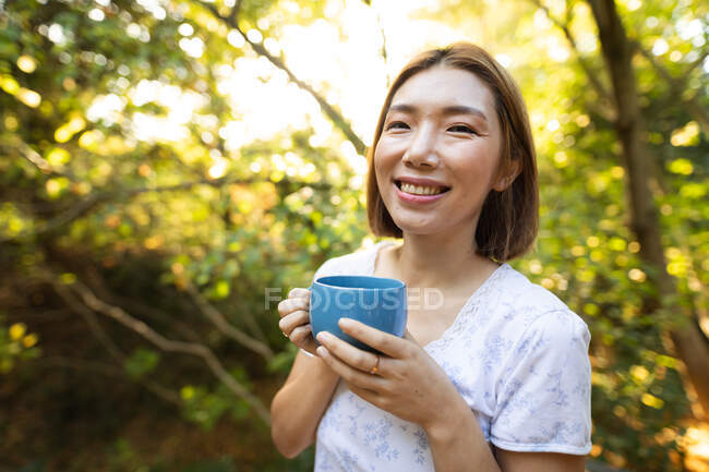 Portrait de femme asiatique souriante en pyjama tenant une tasse de thé debout dans le jardin. à domicile en isolement pendant le confinement en quarantaine. — Photo de stock