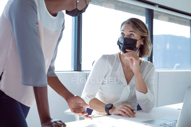 Two diverse smiling businesswomen wearing face masks, working together, using laptop, talking. independent creative business at a modern office during coronavirus covid 19 pandemic. — Stock Photo