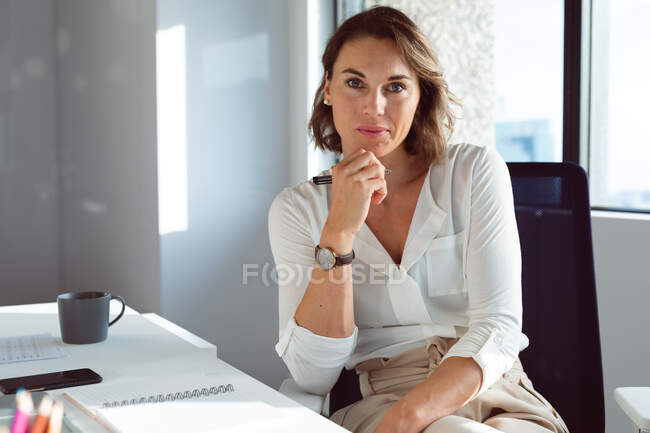 Кавказская деловая женщина сидит за столом, держит ручку на работе. независимый творческий бизнес в современном офисе. — стоковое фото