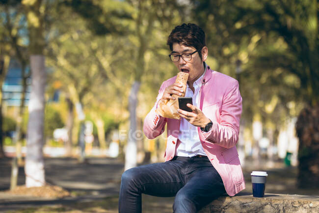 Asiatischer Geschäftsmann, der mit seinem Smartphone Kaffee zum Mitnehmen trinkt und die Straße überquert. Digitaler Nomade im Stadtkonzept unterwegs. — Stockfoto