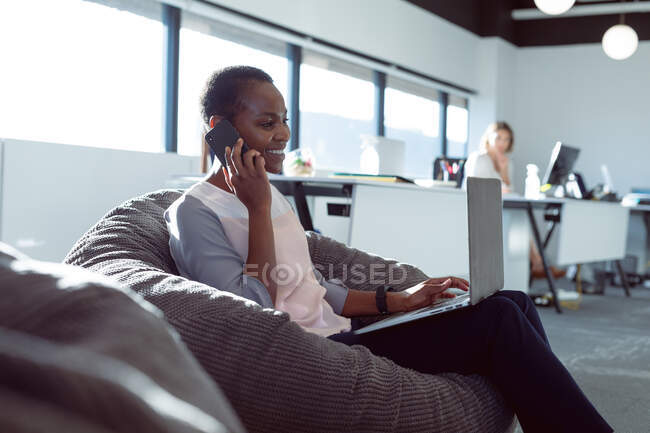 Una donna d'affari afroamericana sorridente seduta in poltrona, che parla con lo smartphone al lavoro. attività creativa indipendente in un ufficio moderno. — Foto stock