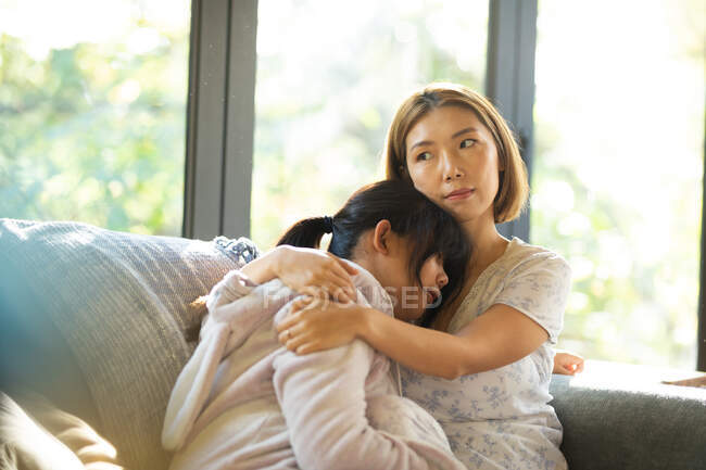 Retrato de una mujer asiática seria con su hija abrazada sentada en un sofá. en casa en aislamiento durante el bloqueo de cuarentena. - foto de stock