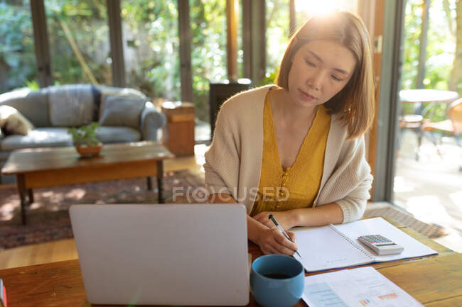 Mulher asiática usando laptop fazendo anotações sentado à mesa, trabalhando em casa. em casa em isolamento durante o confinamento de quarentena. — Fotografia de Stock