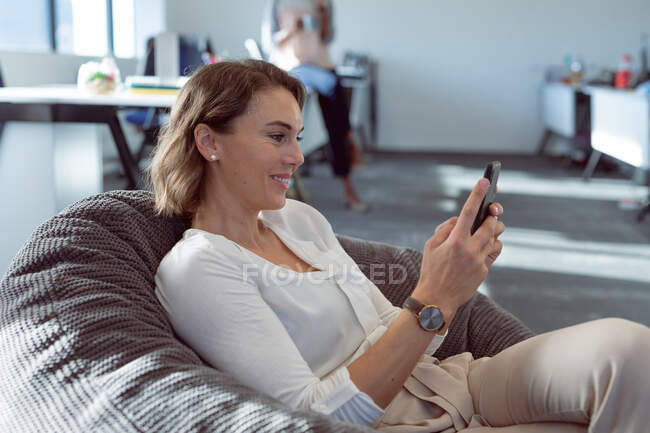 Lächelnde kaukasische Geschäftsfrau, die im Sessel sitzt und ihr Smartphone benutzt. unabhängiges kreatives Geschäft in einem modernen Büro. — Stockfoto