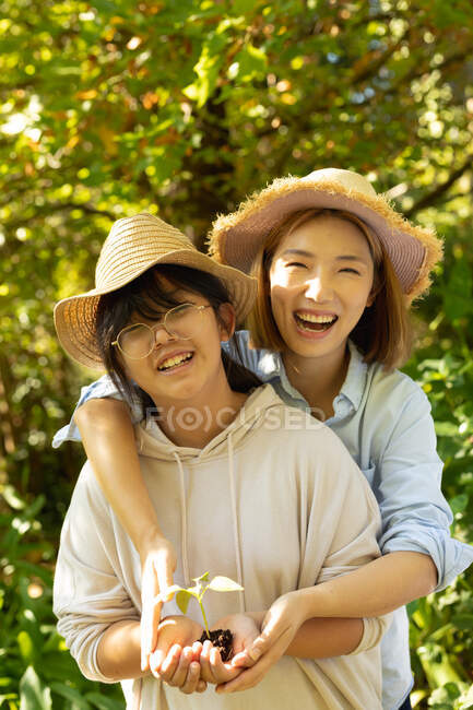 Mujer asiática sonriente con su hija usando sombreros de paja y sosteniendo la planta en el jardín. en casa en aislamiento durante el bloqueo de cuarentena. - foto de stock