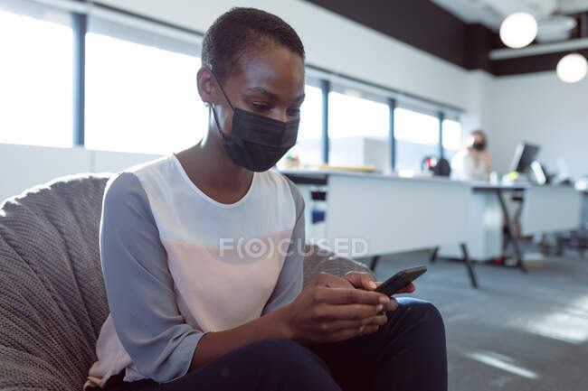 Smiling african american businesswoman sitting in armchair, wearing face mask, using smartphone. independent creative business at a modern office during coronavirus covid 19 pandemic. — Stock Photo