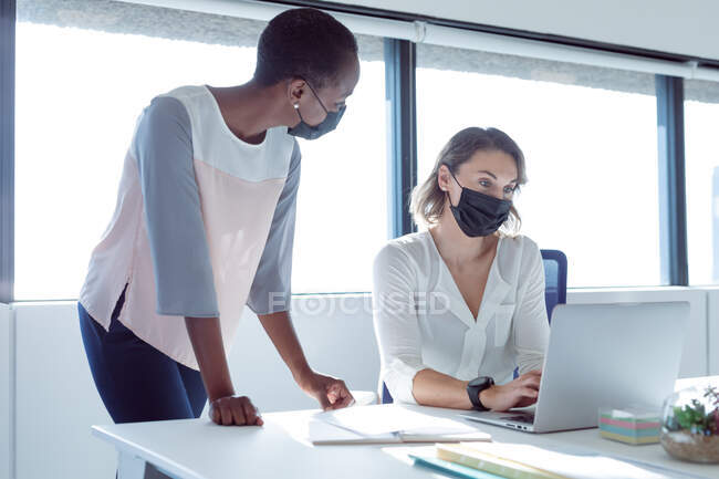 Duas empresárias sorrindo diversas usando máscaras faciais, trabalhando juntas, usando laptop, conversando. negócio criativo independente em um escritório moderno durante coronavírus covid 19 pandemia. — Fotografia de Stock