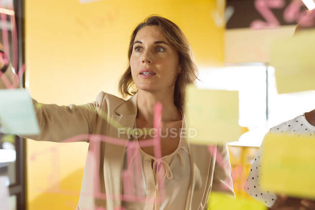 Femme d'affaires caucasienne prenant des notes et ajoutant des post-ins sur un tableau transparent. entreprise créative indépendante dans un bureau moderne. — Photo de stock
