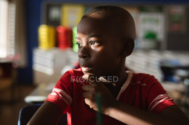 Ragazzo afroamericano che guarda fuori dalla finestra mentre si siede sulla scrivania in classe alle elementari. concetto di scuola e istruzione — Foto stock