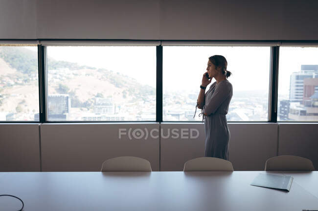 Mujer de negocios caucásica de pie en la ventana, hablando por teléfono inteligente en el trabajo. negocio creativo independiente en una oficina moderna. - foto de stock