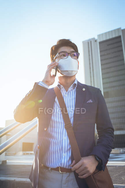 Asian businessman wearing face mask using smartphone in city street. digital nomad out and about in city during covid 19 pandemic concept. — Stock Photo