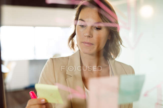Donna d'affari caucasica che prende appunti e aggiunge post-in sul tabellone trasparente. attività creativa indipendente in un ufficio moderno. — Foto stock
