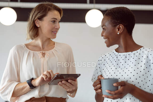Duas empresárias diversas conversando, sorrindo, segurando tablet e café. negócio criativo independente em um escritório moderno. — Fotografia de Stock