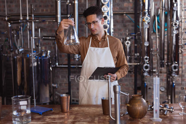 Trabalhador caucasiano vestindo avental segurando um frasco e uma prancheta na destilaria de gin. conceito de produção e filtração de álcool — Fotografia de Stock