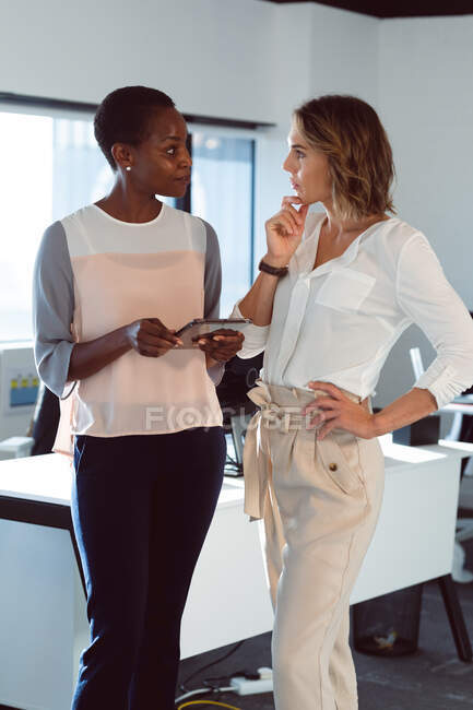 Dos empresarias diversas de pie, usando tableta y hablando en el trabajo. negocio creativo independiente en una oficina moderna. - foto de stock