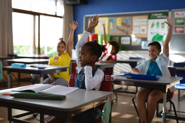 Grupo de estudantes diversos levantando as mãos na classe no ensino fundamental. conceito de escola e educação — Fotografia de Stock
