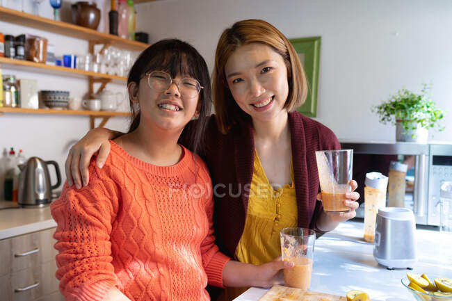 Retrato de mãe asiática sorridente e filha com smoothie caseiro na cozinha. em casa em isolamento durante o confinamento de quarentena. — Fotografia de Stock