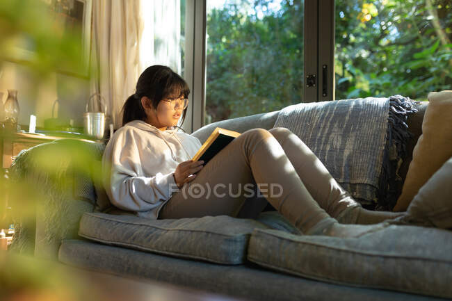 Sorrindo menina asiática em óculos lendo um livro e sentado no sofá. em casa em isolamento durante o confinamento de quarentena. — Fotografia de Stock