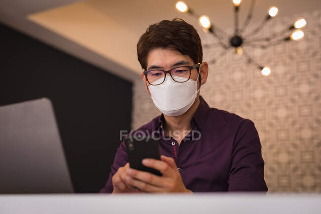 Asian businessman wearing face mask using smartphone and laptop in hotel lobby. digital nomad out and about in city during covid 19 pandemic concept. — Stock Photo