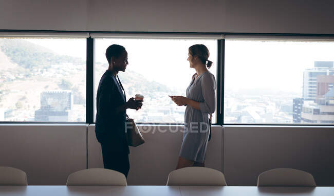 Dos empresarias diversas de pie en la ventana, hablando en el trabajo. negocio creativo independiente en una oficina moderna. - foto de stock