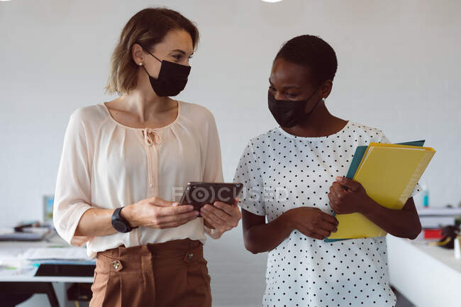 Zwei unterschiedliche Geschäftsfrauen mit Mundschutz, Tablet und Dokumenten in der Hand, unterhalten sich. Selbstständiges kreatives Geschäft in einem modernen Büro während der Coronavirus-Pandemie 19. — Stockfoto