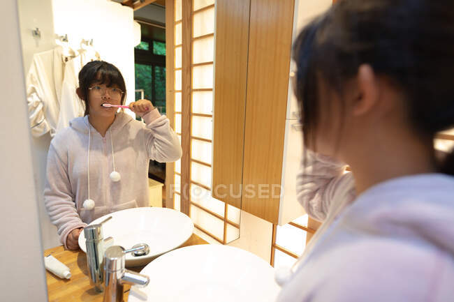 Menina asiática em óculos escovando os dentes no banheiro. em casa em isolamento durante o confinamento de quarentena. — Fotografia de Stock