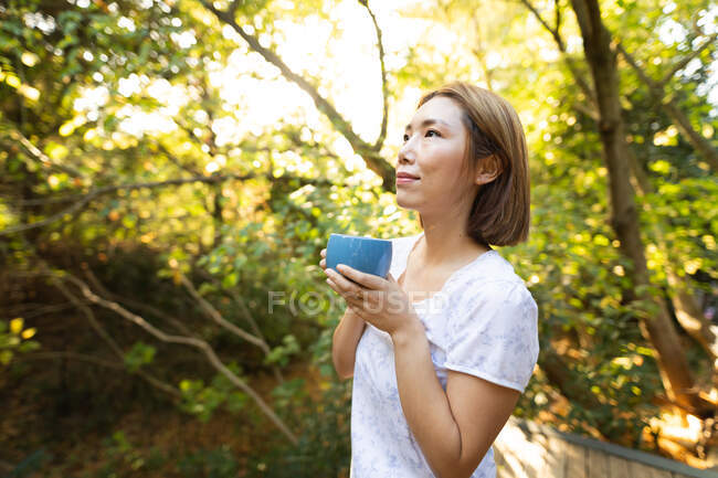 Porträt einer lächelnden asiatischen Frau im Pyjama mit Teetasse im Garten. Zuhause in Isolation während der Quarantäne. — Stockfoto
