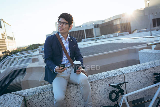 Asiatischer Geschäftsmann mit Smartphone und Kaffee zum Mitnehmen sitzt an seinem Fahrrad in der Stadtstraße. Digitaler Nomade im Stadtkonzept unterwegs. — Stockfoto