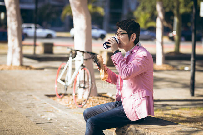 Asian businessman using smartphone drinking takeaway coffee sitting on wall in city. digital nomad out and about in city concept. — Stock Photo