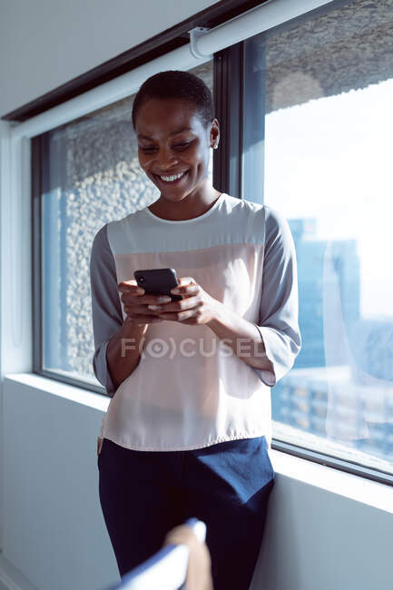 Sorrindo afro-americana empresária de pé na janela, usando smartphone. negócio criativo independente em um escritório moderno. — Fotografia de Stock